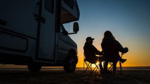A couple sit outside their RV Camper at sunset at the lake.